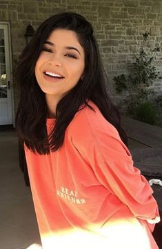 a woman with long dark hair wearing an orange shirt and smiling at the camera while sitting on a porch