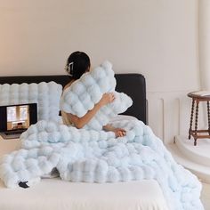 a woman sitting on top of a bed covered in fluffy blue blankets and holding a pillow