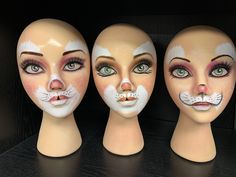 three mannequin heads with painted faces and cats on their faces, one has green eyes