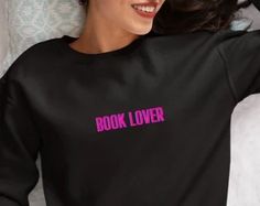 a woman laying on top of a bed wearing a black shirt with pink letters reading book lover