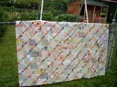 a quilt hanging on a clothes line in the grass near a fence with a house behind it