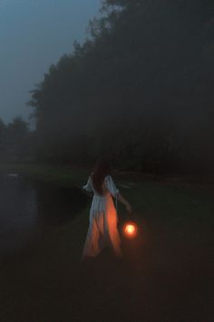 a woman holding a lantern in the dark by a body of water with trees behind her