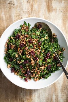 a white bowl filled with greens and nuts on top of a wooden table next to a spoon