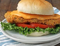 a chicken sandwich with lettuce and tomatoes on a white plate next to a blue and white towel