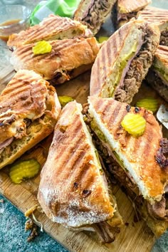 a wooden cutting board topped with cut in half sandwiches on top of each other next to pickles