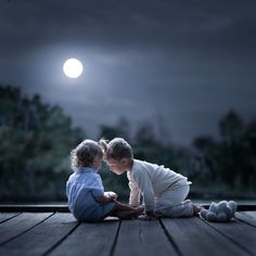 two children sitting on the ground in front of a full moon at night with their faces touching each other