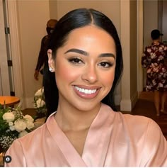 a woman in a pink robe smiling at the camera with flowers on the table behind her