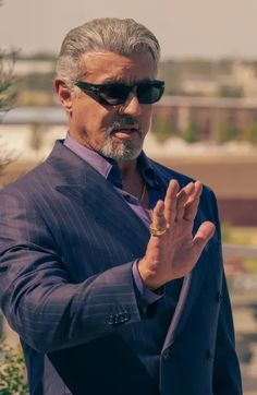 a man in a suit and sunglasses standing next to a tree with his hands together