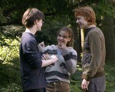 three young men standing next to each other in front of some trees and bushes, talking