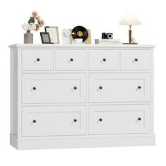 a white dresser with many drawers and pictures on it's top, next to a lamp