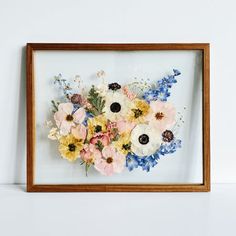 an arrangement of flowers is framed in a wooden frame on a white wall with a shelf