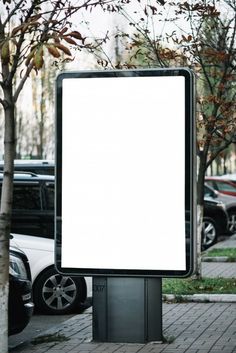 an empty billboard stands on the sidewalk in front of parked cars and tree's