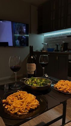 two plates of food on a table with wine and a tv in the background at night