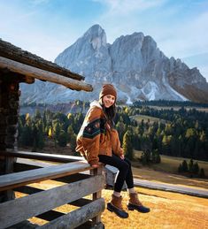 a woman sitting on top of a wooden fence in front of a mountainside area