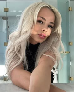 a woman with long blonde hair sitting in front of a bathroom mirror looking at the camera