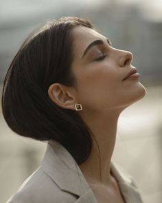 a woman with her eyes closed looking up to the side, wearing a white blazer and gold earrings