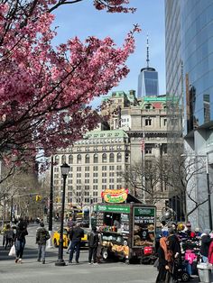 many people are walking on the street near some buildings and trees with pink flowers in bloom