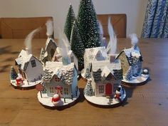 three small houses with snow on the roofs and trees in front of them, sitting on a wooden table