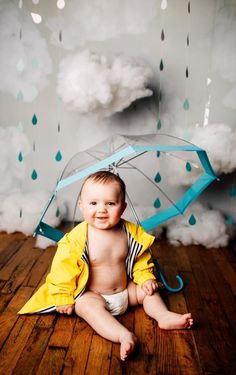 a baby sitting on the floor with an umbrella in front of him and clouds behind it