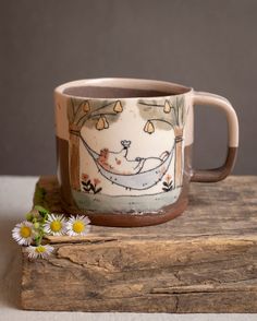 a ceramic mug with a cat sleeping in a hammock on top of a piece of wood