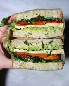 a person holding a sandwich in their hand with lettuce, tomato and avocado on it