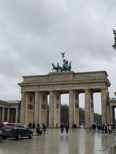 people are walking around in front of an arch with statues on it and cars parked nearby