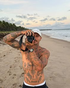 a tattooed man taking a photo on the beach with his camera in front of him