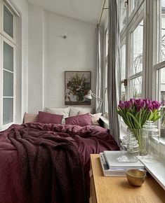 a bed with purple sheets and flowers in vases on the table next to it