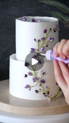 someone is decorating a cake with purple flowers on the top and bottom tiers