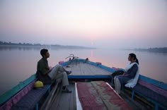 two people are sitting on the back of a boat at sunset or dawn, looking out over the water