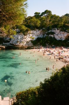 many people are swimming in the clear blue water on a beach with cliffs behind them