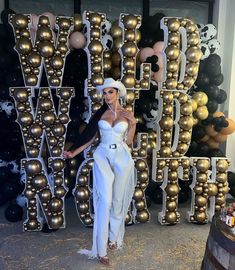 a woman standing in front of a giant letter with gold balls behind her and wearing a white hat