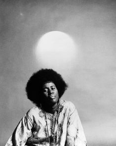 a black and white photo of a man with an afro sitting on a stool in front of the sun