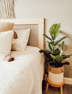 a bed with white sheets and pillows next to a potted plant on a wooden stand