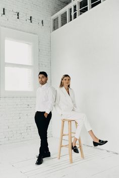 a man and woman sitting on stools in an empty room with brick wall behind them