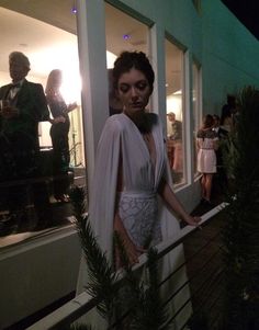 a woman in white dress standing on balcony next to trees and people looking out the window