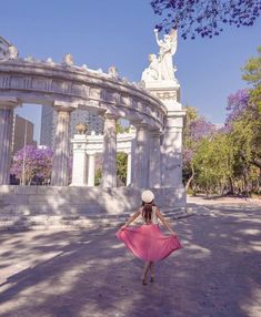 a woman in a pink dress and white hat is walking through the park with her hands behind her back