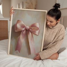 a woman sitting on a bed holding up a painting with a pink bow on it