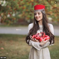 a woman in a white dress is holding some apples and wearing a red headdress