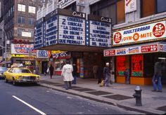 people are walking on the sidewalk in front of a movie theater with neon signs and advertisements