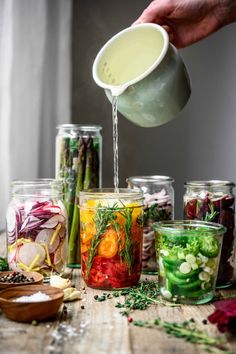 a person pours water into jars filled with different types of vegetables and herbs on a wooden table