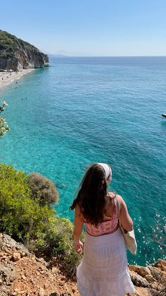 a woman in a white dress is looking out at the ocean from a cliff side