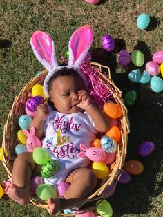 a baby wearing bunny ears sitting in a basket with easter eggs around her and on the ground