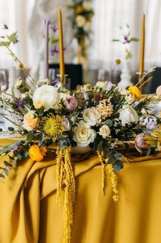 an arrangement of flowers and greenery on a yellow table cloth with candles in the background