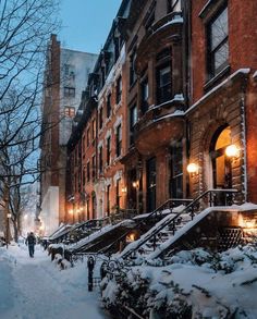 people walking down a snow covered street in the city at night with buildings and lights on