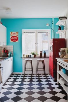 a black and white checkered floor in a small kitchen with blue walls, an old fashioned refrigerator and two stools