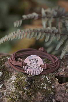 a leather bracelet with a metal disc that says enjoy the journey on it sitting on top of a mossy rock