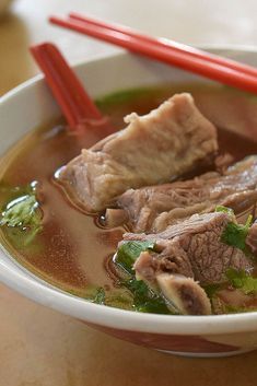 a bowl of soup with meat and broccoli in it, next to two red chopsticks