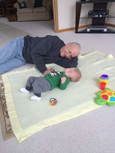 an older man laying on top of a blanket next to a baby in a green shirt