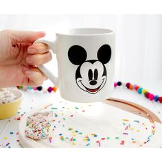 a person holding a mickey mouse coffee mug with sprinkles on the table